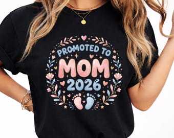 May include: Black t-shirt with a colorful graphic that reads "Promoted to MOM 2026." The design includes floral elements, hearts, stars, and baby footprints. The text is in a playful font.