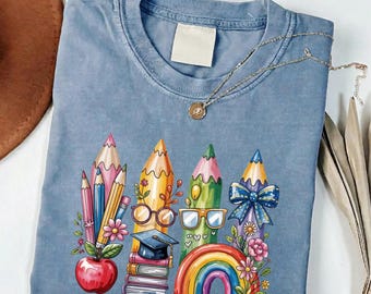 May include: A blue jean colored t-shirt featuring a graphic print of colorful pencils, a red apple, a stack of books with a graduation cap, eyeglasses, a rainbow, and floral accents. A delicate gold-toned chain necklace rests on the neckline.