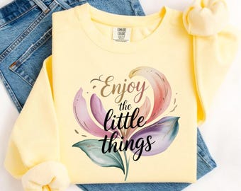 May include: A pale yellow long-sleeve sweatshirt featuring a graphic of a multi-colored flower with petals in shades of pink, purple, and teal. The text "Enjoy the little things" is printed in black cursive lettering over the center of the flower.