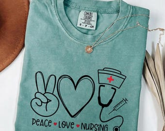 May include: A sage green t-shirt featuring a black graphic design with a peace sign, a heart, a nurse cap with a red cross, a stethoscope, and a syringe. The text reads PEACE LOVE NURSING with small red hearts between the words. The shirt has a Comfort Colors label.