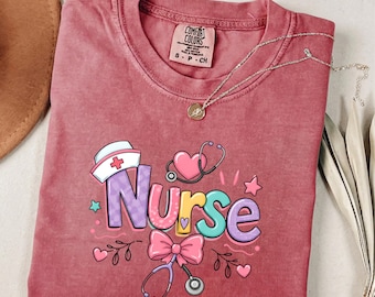 May include: A dusty rose short-sleeve t-shirt featuring the word Nurse in colorful, patterned bubble letters. The graphic includes a white nurse cap, a pink heart, a stethoscope, and a pink bow. The shirt has a Comfort Colors brand label.