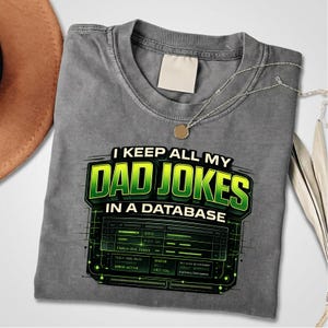 Op de afbeelding: Grijze t-shirt met de tekst "I KEEP ALL MY DAD JOKES IN A DATABASE" in groen en zwart. De afbeelding bevat een computerdatabase-ontwerp. Een zilveren ketting met een ronde hanger ligt op het shirt.
