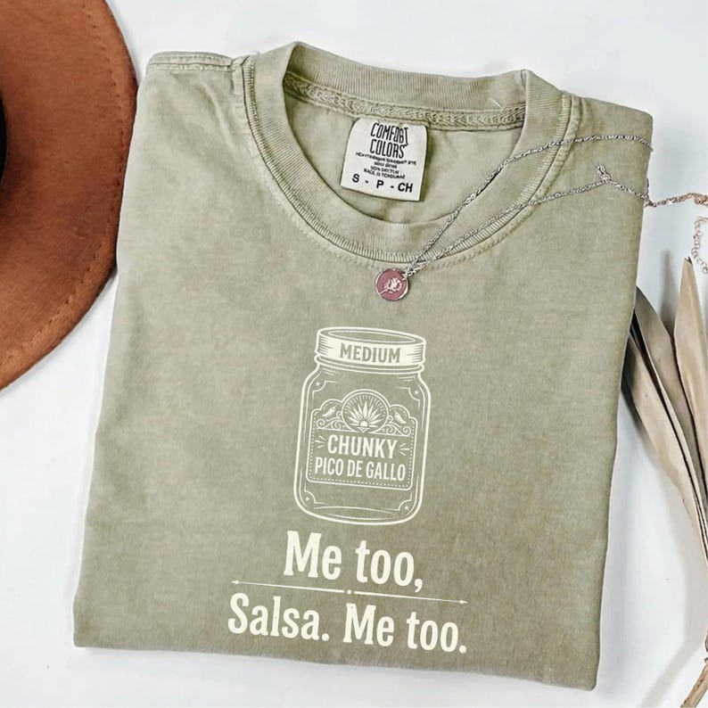 May include: An olive green t-shirt with the text "Me too, Salsa. Me too." and a graphic of a jar labeled "Chunky Pico de Gallo." A silver necklace with a pink pendant rests on the shirt.