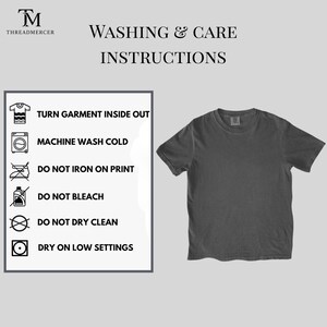 May include: A dark gray t-shirt next to a panel with washing and care instructions. The instructions include turning the garment inside out, machine washing cold, and avoiding ironing on the print, bleaching, and dry cleaning. The shirt has a crew neck.