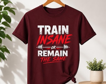 May include: A maroon short-sleeved t-shirt featuring white and red text that reads "TRAIN INSANE OR REMAIN THE SAME" with a graphic of a barbell centered between the words. The shirt is displayed on a wooden hanger against a plain background.
