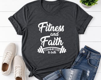 May include: A dark charcoal gray short-sleeve t-shirt featuring the white text Fitness and Faith COMMITTED to both above a barbell graphic. The shirt is displayed alongside a pair of light beige sneakers and blue denim jeans with distressed details.