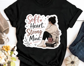May include: A black short-sleeved t-shirt featuring a graphic illustration of a person sitting with books, a mug, and a candle. The text reads Soft Heart, Strong Mind. The design includes decorative hearts, stars, and a small floral arrangement.