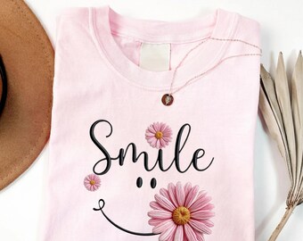 May include: A light pink short-sleeved t-shirt featuring the word Smile in black cursive script. The design includes two pink daisy flowers with yellow centers positioned as eyes and a large pink daisy forming part of a black curved smile line.