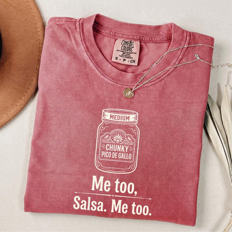 May include: A red-pink t-shirt with the text "Me too, Salsa. Me too." and a graphic of a jar labeled "Chunky Pico de Gallo." The shirt has a crew neck and short sleeves. A silver necklace with a pendant is on the shirt.