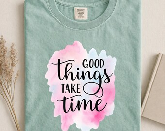 May include: A sage green short-sleeve t-shirt featuring a central watercolor-style graphic in shades of pink and light blue. The black text printed over the graphic reads "GOOD things TAKE time" in a mix of serif and script fonts.
