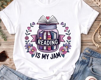 May include: A white short-sleeve t-shirt featuring a graphic of a glass jar filled with colorful books, topped with an open book and a small heart. The text "READING IS MY JAM" is printed below the jar, framed by pink and purple floral vines.