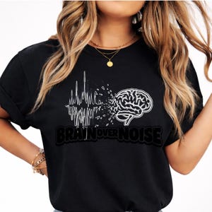 May include: Black t-shirt with a white graphic. The design shows a brain and a sound wave, with the text "BRAIN OVER NOISE". The shirt is made of a soft material. The text is in a bold font.