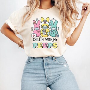 May include: Cream-colored t-shirt featuring a graphic of three cartoon bunnies wearing face masks and medical accessories. The text "CHILLIN' WITH MY PEEPS" is below the bunnies in colorful letters. The shirt is worn by a person.