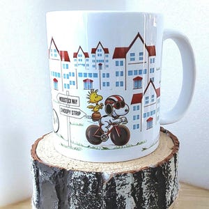 May include: White ceramic mug featuring a cartoon illustration of Snoopy riding a bicycle with Woodstock. The mug has a cityscape design with red-roofed buildings and street signs that read "Woodstock Way" and "Snoopy Strip."