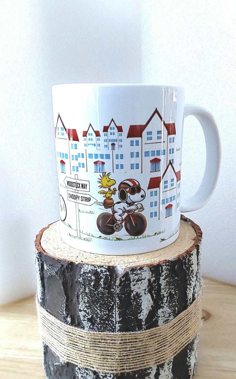 May include: White ceramic mug featuring a cartoon illustration of Snoopy riding a bicycle with Woodstock. The mug has a cityscape design with red-roofed buildings and street signs that read "Woodstock Way" and "Snoopy Strip."