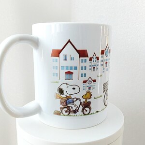May include: White ceramic mug featuring a cartoon Snoopy character riding a bicycle with Woodstock. The mug has a wrap-around design of buildings with red roofs and blue windows. The mug has a large, curved handle.