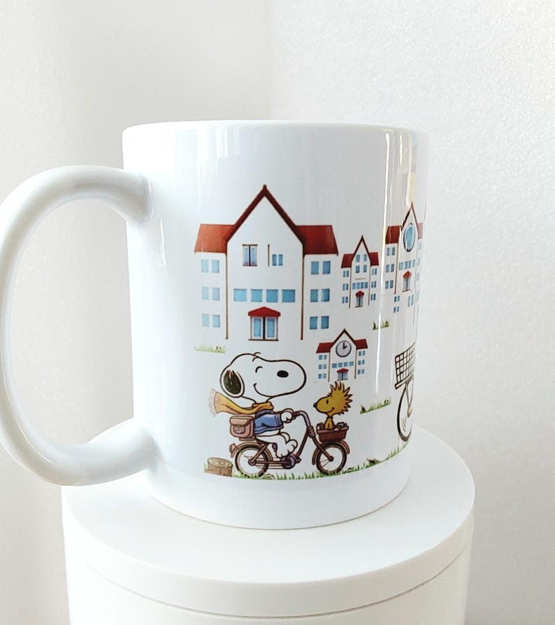 May include: White ceramic mug featuring a cartoon Snoopy character riding a bicycle with Woodstock. The mug has a wrap-around design of buildings with red roofs and blue windows. The mug has a large, curved handle.