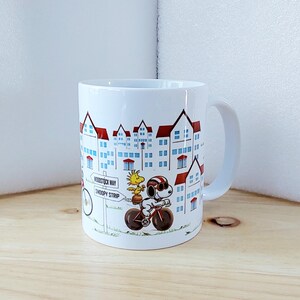May include: White ceramic mug featuring a cartoon illustration of Snoopy and Woodstock riding a bicycle in front of a townscape. The mug has a curved handle and the words "Woodstock Way" and "Snoopy Strip" are visible.