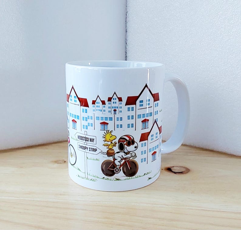 May include: White ceramic mug featuring a cartoon illustration of Snoopy and Woodstock riding a bicycle in front of a townscape. The mug has a curved handle and the words "Woodstock Way" and "Snoopy Strip" are visible.