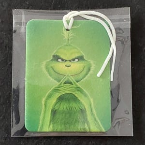 May include: A rectangular air freshener featuring a green, furry character with a mischievous expression and hands pressed together, printed on a light green background. The item is enclosed in a clear plastic sleeve and includes a white elastic string.