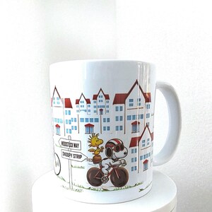 May include: White ceramic mug featuring a cartoon illustration of Snoopy riding a bicycle with Woodstock. The design includes a cityscape with red-roofed buildings and a sign that reads "Woodstock Way" and "Snoopy Strip".