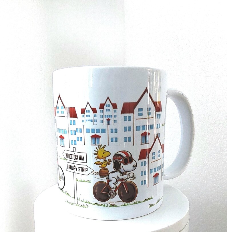 May include: White ceramic mug featuring a cartoon illustration of Snoopy riding a bicycle with Woodstock. The design includes a cityscape with red-roofed buildings and a sign that reads "Woodstock Way" and "Snoopy Strip".