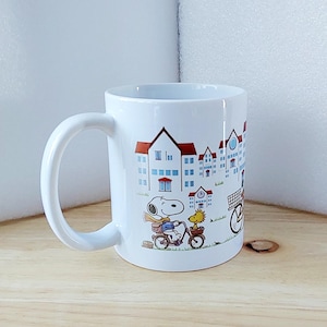May include: White ceramic mug featuring a colorful illustration of Snoopy and Woodstock riding bicycles in front of a townscape. The mug has a curved handle and a glossy finish, perfect for coffee or tea.