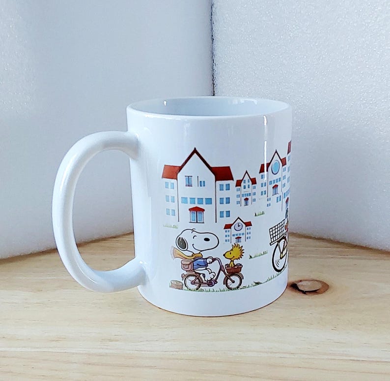 May include: White ceramic mug featuring a colorful illustration of Snoopy and Woodstock riding bicycles in front of a townscape. The mug has a curved handle and a glossy finish, perfect for coffee or tea.