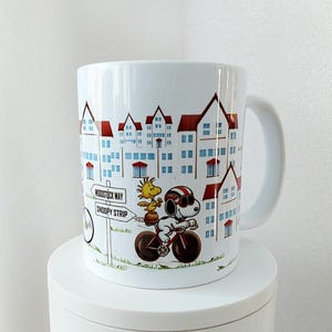 May include: White ceramic mug featuring a cartoon illustration of Snoopy riding a bicycle with Woodstock. The mug has a cityscape design with red-roofed buildings and the text "Woodstock Way" and "Snoopy Strip".