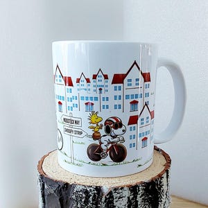 May include: White ceramic mug featuring a cartoon illustration of Snoopy riding a bicycle with Woodstock. The mug has a cityscape design with red-roofed buildings and the text "Woodstock Way" and "Snoopy Strip".
