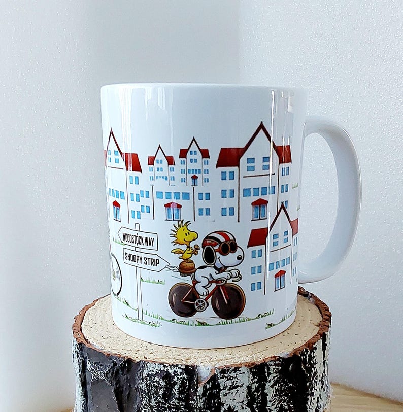 May include: White ceramic mug featuring a cartoon illustration of Snoopy riding a bicycle with Woodstock. The mug has a cityscape design with red-roofed buildings and the text "Woodstock Way" and "Snoopy Strip".