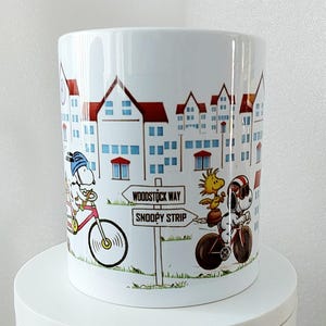 May include: White ceramic mug featuring a cartoon illustration of Snoopy and Woodstock riding bicycles in a town setting. The mug has a cityscape design with red-roofed buildings and a signpost that reads "Woodstock Way" and "Snoopy Strip."