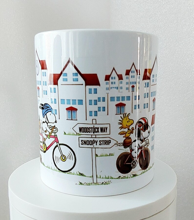 May include: White ceramic mug featuring a cartoon illustration of Snoopy and Woodstock riding bicycles in a town setting. The mug has a cityscape design with red-roofed buildings and a signpost that reads "Woodstock Way" and "Snoopy Strip."