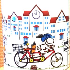 May include: White ceramic mug featuring Snoopy and Woodstock riding a tandem bicycle in front of a town scene. The red bicycle has a basket and is set against a backdrop of buildings with red roofs and blue windows.