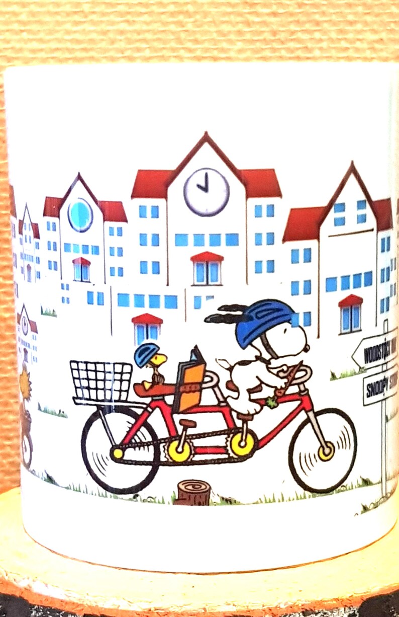 May include: White ceramic mug featuring Snoopy and Woodstock riding a tandem bicycle in front of a town scene. The red bicycle has a basket and is set against a backdrop of buildings with red roofs and blue windows.