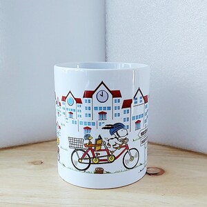 May include: White ceramic mug featuring a whimsical illustration of a dog and a duck riding a tandem bicycle in front of a townscape. The buildings have red roofs and blue windows. The mug has a black rim and a curved handle.