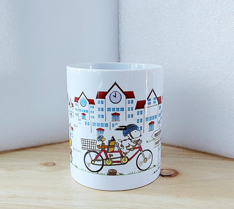 May include: White ceramic mug featuring a whimsical illustration of a dog and a duck riding a tandem bicycle in front of a townscape. The buildings have red roofs and blue windows. The mug has a black rim and a curved handle.