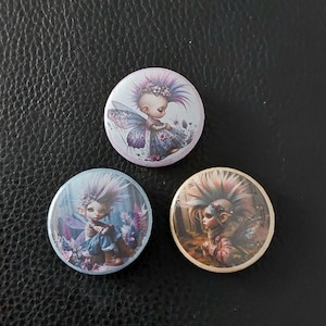 May include: Three circular pin-back badges featuring illustrations of fairy-like figures with spiky hair and wings. The top badge has a light purple background, the bottom left has a blue background, and the bottom right has a warm brown background.