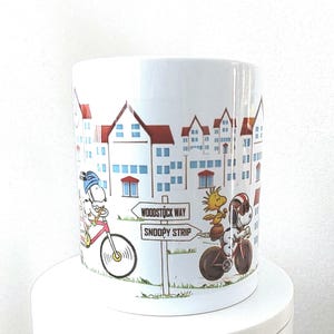 May include: White ceramic mug featuring a cartoon illustration of Snoopy and Woodstock riding bicycles. The mug has a cityscape design with red-roofed buildings and street signs that read "Woodstock Way" and "Snoopy Strip."