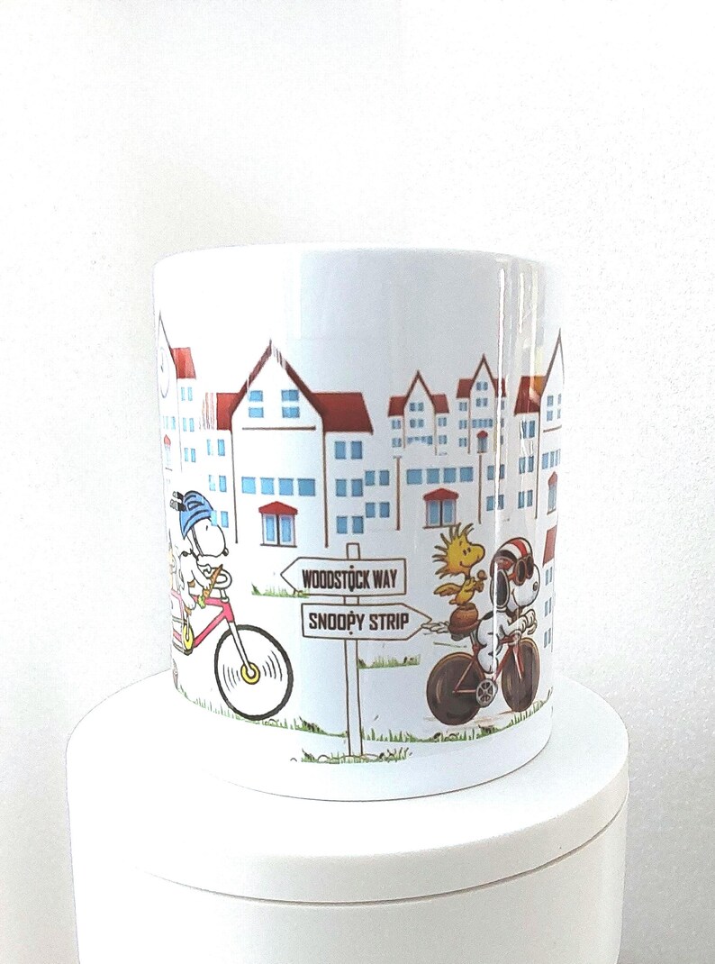 May include: White ceramic mug featuring a cartoon illustration of Snoopy and Woodstock riding bicycles. The mug has a cityscape design with red-roofed buildings and street signs that read "Woodstock Way" and "Snoopy Strip."