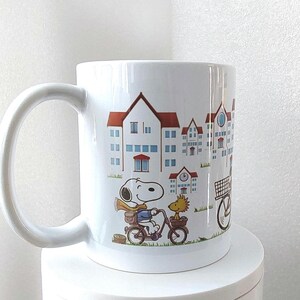 May include: White ceramic mug featuring a cartoon illustration of Snoopy riding a bicycle with Woodstock. The mug has a cityscape design with blue and red buildings. The mug has a large, curved handle.