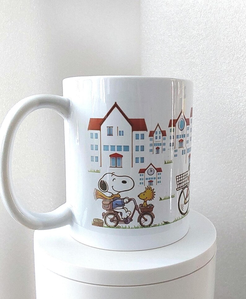 May include: White ceramic mug featuring a cartoon illustration of Snoopy riding a bicycle with Woodstock. The mug has a cityscape design with blue and red buildings. The mug has a large, curved handle.