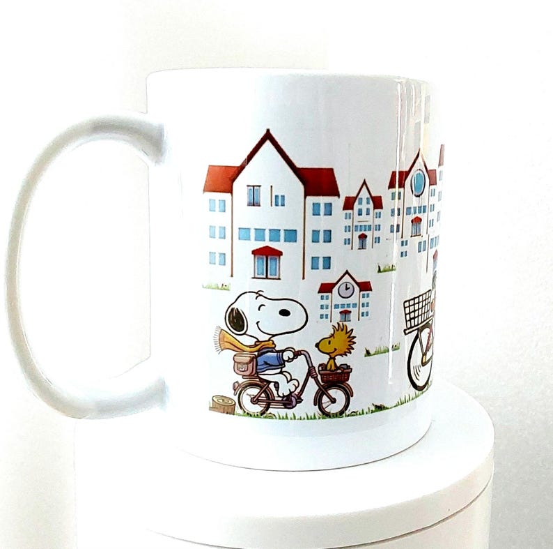 May include: White ceramic mug featuring a cartoon illustration of Snoopy and Woodstock riding a bicycle through a town with red-roofed buildings. The mug has a large, curved handle.