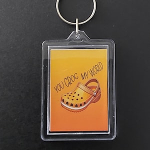 May include: A rectangular clear acrylic keyring featuring an orange gradient background with an illustration of a yellow clog shoe and the black text "YOU CROC MY WORLD" printed above the shoe. The keyring includes a silver metal ring.