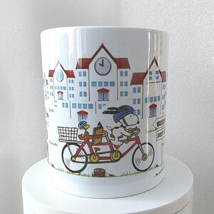 May include: White ceramic mug featuring a cartoon illustration of Snoopy and Woodstock riding a tandem bicycle through a town. The buildings have red roofs and blue windows. The mug has a glossy finish.