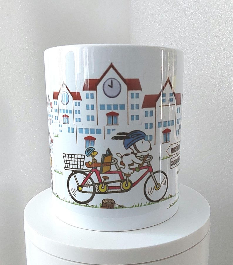 May include: White ceramic mug featuring a cartoon illustration of Snoopy and Woodstock riding a tandem bicycle through a town. The buildings have red roofs and blue windows. The mug has a glossy finish.