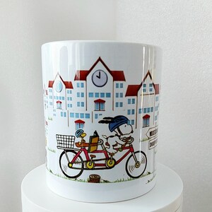 May include: White ceramic mug featuring a cartoon illustration of Snoopy riding a tandem bicycle with Woodstock. The mug has a red and blue color scheme with a town scene background. The mug is on a white surface.