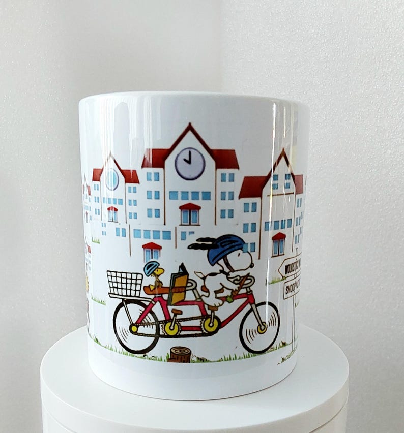 May include: White ceramic mug featuring a cartoon illustration of Snoopy riding a tandem bicycle with Woodstock. The mug has a red and blue color scheme with a town scene background. The mug is on a white surface.