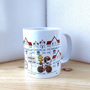 May include: White ceramic mug featuring a cartoon illustration of Snoopy riding a bicycle with Woodstock. The mug has a cityscape design with red-roofed buildings and a sign that reads "Woodstock Way Snoopy Strip."
