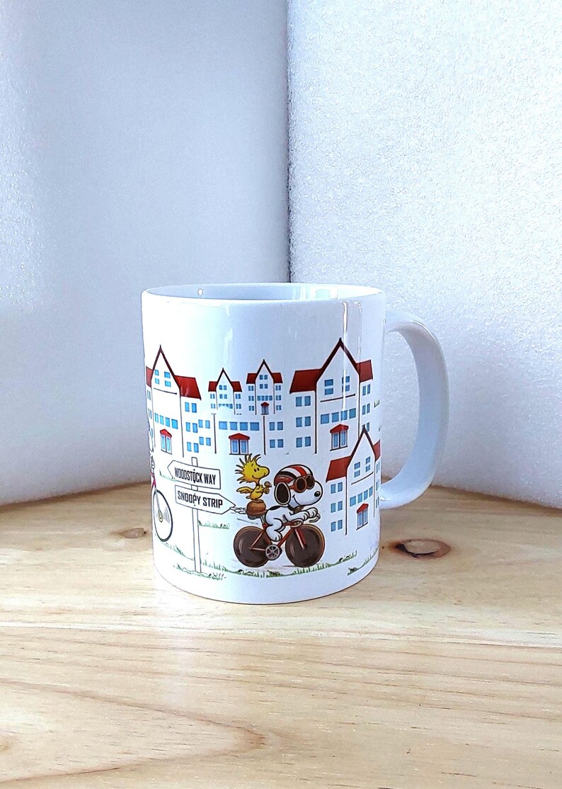 May include: White ceramic mug featuring a cartoon illustration of Snoopy riding a bicycle with Woodstock. The mug has a cityscape design with red-roofed buildings and a sign that reads "Woodstock Way Snoopy Strip."